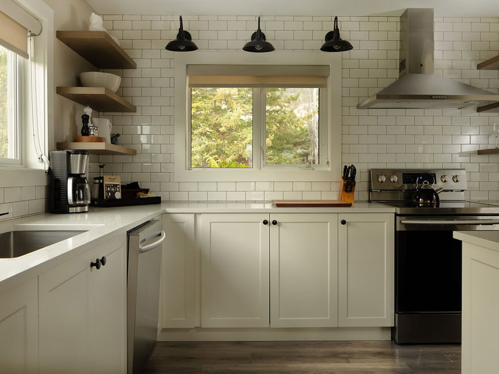 A modern kitchen with white cabinets and subway tile backsplash. Stainless steel appliances, including a stove and dishwasher, are present. Open shelves with dishes and a window offering a view of trees enhance the space.