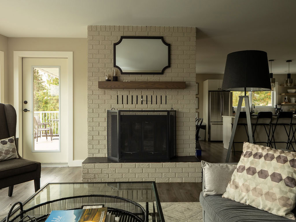 A cozy living room with a brick fireplace and a wooden mantel, above which hangs an oval mirror. A glass door to the left opens to a balcony. The room features neutral tones, a gray sofa, a patterned cushion, and a black floor lamp.