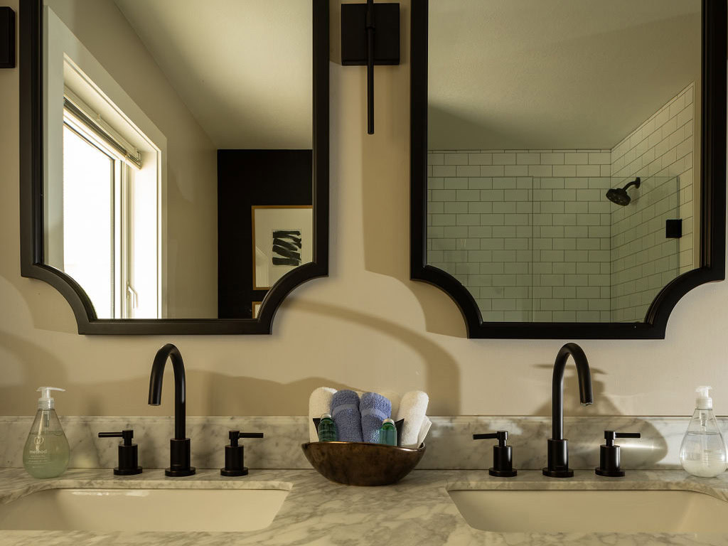 A modern bathroom features two black-framed mirrors above a marble countertop with double sinks. Black fixtures complement the decor. A wooden bowl holds rolled towels, and a shower with white tiles is visible in the background.