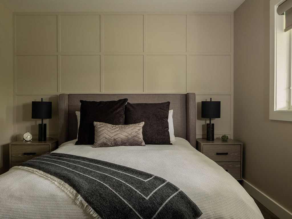 A modern bedroom featuring a neatly made bed with a gray upholstered headboard, accent pillows, and a striped throw blanket. Two black lamps sit on wooden nightstands beside the bed, against a paneled beige wall. Natural light filters through a nearby window.