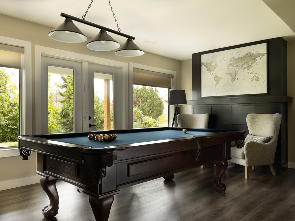 A cozy room with a pool table centered under a three-light fixture. Two armchairs sit by a large window with a view of trees. A map hangs on the wall, and natural light fills the space, highlighting the wood flooring.