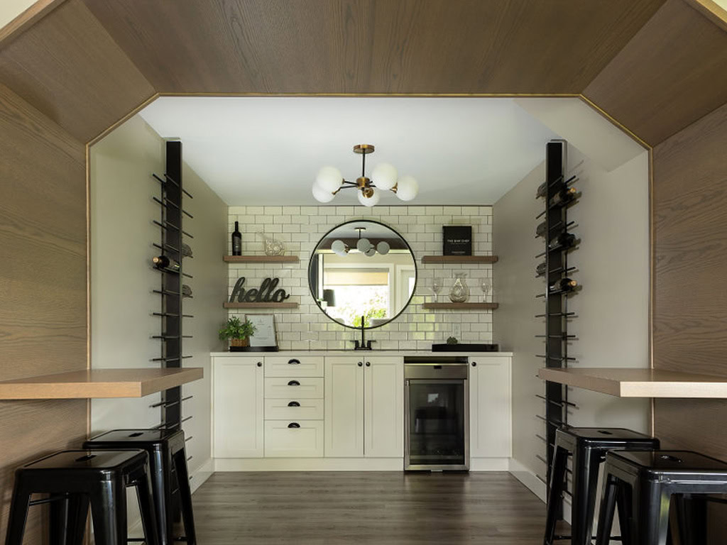 Modern kitchen with white cabinets, a round mirror, and a black and white backsplash. Two tables with black stools flank the sides. A central light fixture hangs from the ceiling, and wine racks are mounted on the walls.