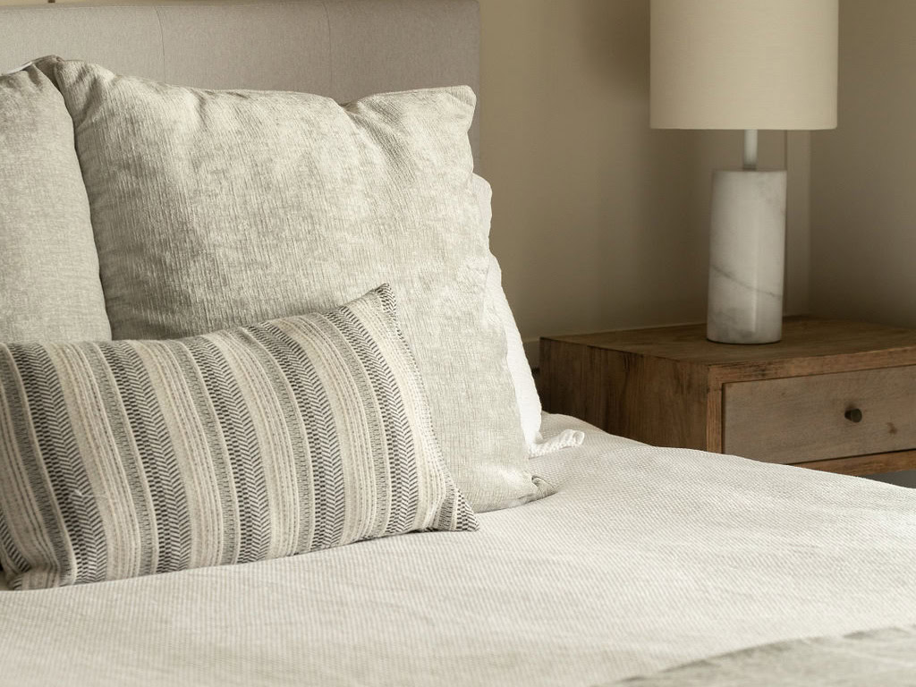 A cozy bedroom scene with a neatly made bed featuring light-colored pillows. A wooden nightstand sits beside the bed, topped with a marble-textured lamp with a white shade. The overall color palette is neutral and calming.