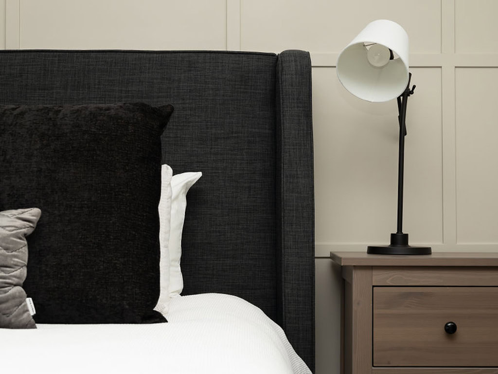 A cozy bedroom corner featuring a dark upholstered headboard with white and dark pillows. A wooden nightstand is beside the bed, topped with a modern black and white lamp against a light paneled wall.