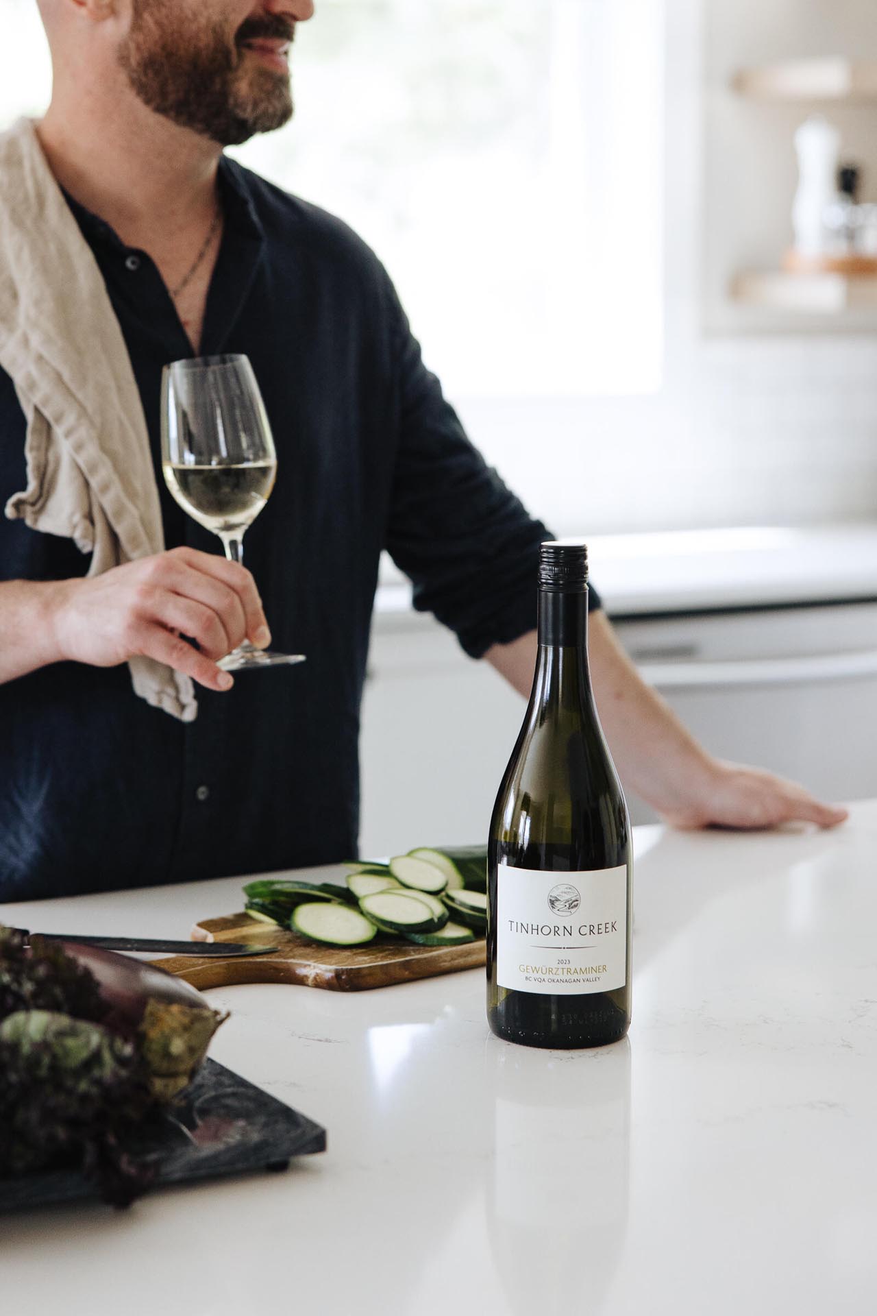 A person in a black shirt holds a glass of white wine in a kitchen. A bottle of wine labeled "Tinhorn Creek" is on the counter, with sliced cucumbers and lettuce nearby.