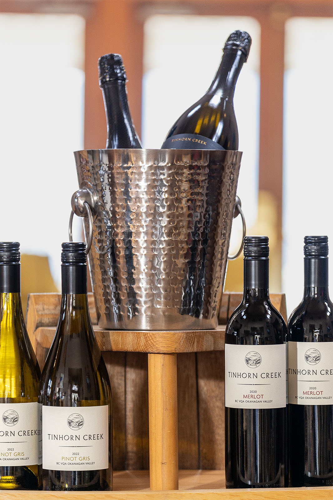 A decorative metal ice bucket holds two bottles of sparkling wine. In front, four bottles of Tinhorn Creek wine—a mix of Pinot Gris and Merlot—are displayed on a wooden surface.