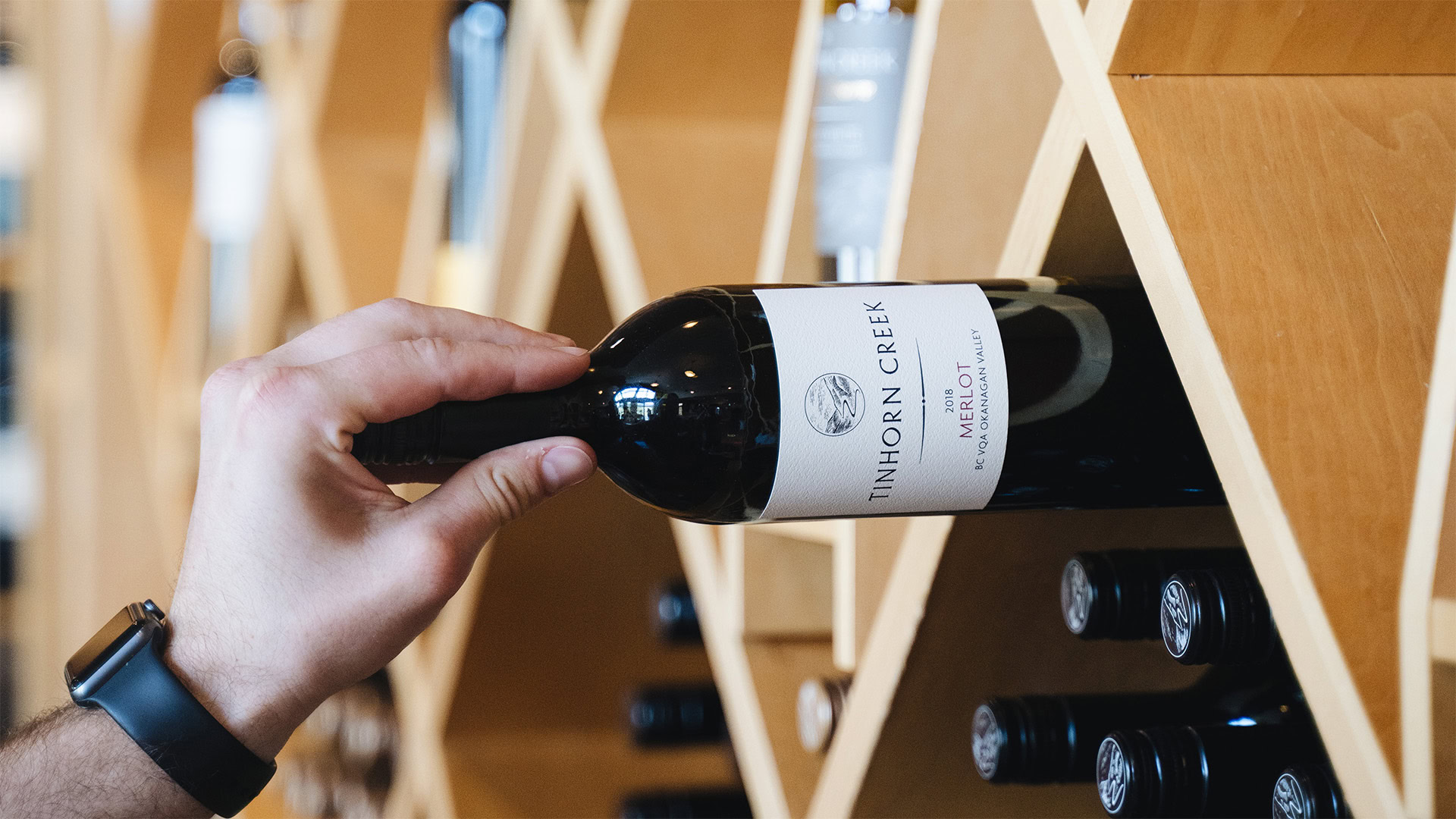 A person wearing a smartwatch retrieves a bottle of Tinhorn Creek red wine from a wooden, diamond-shaped wine rack. Bottles are stored horizontally, and the setting appears to be a wine cellar or store.