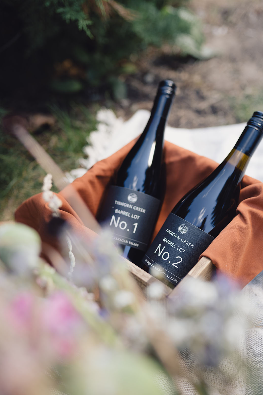 Two wine bottles labeled "Barrel Lot No. 1" and "Barrel Lot No. 2" are placed in a wooden crate lined with a brown cloth. The crate sits outdoors on a white blanket, with blurred flowers in the foreground and greenery in the background.