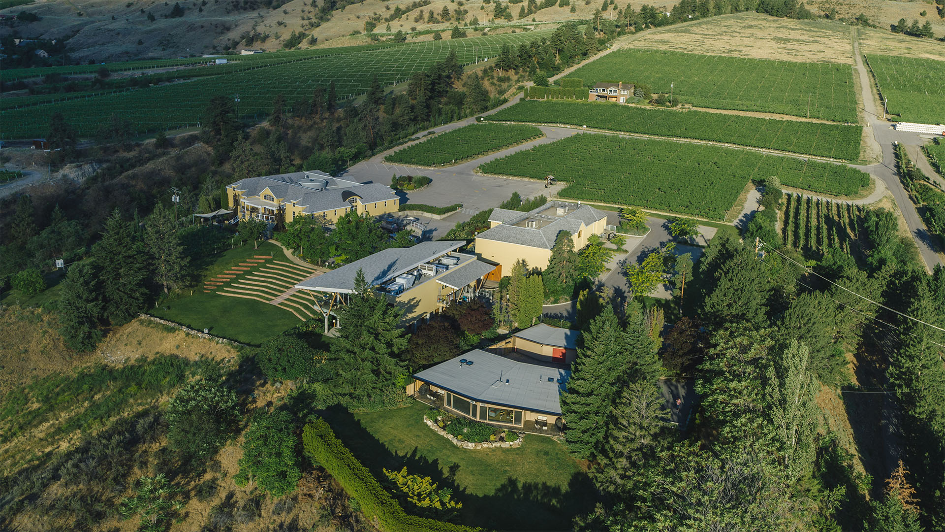 Aerial view of a vineyard estate with multiple buildings surrounded by lush green fields and trees. The landscape includes well-maintained gardens, pathways, and a backdrop of rolling hills under a clear sky.