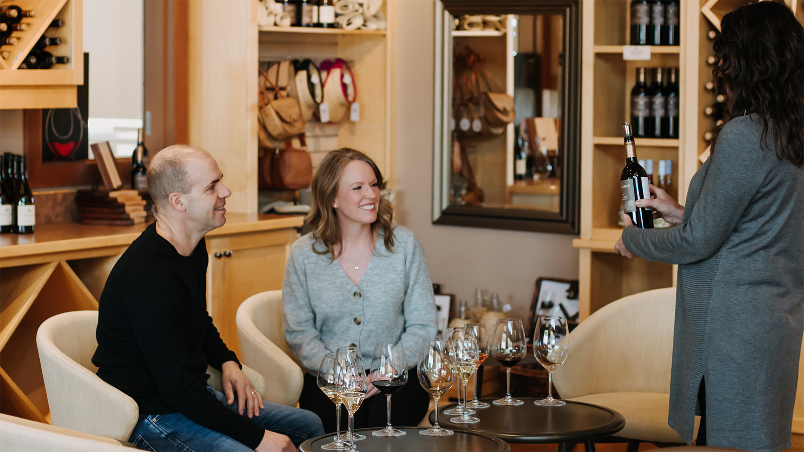 A woman is presenting a wine bottle to a seated couple in a cozy wine shop. The couple is smiling, surrounded by wine glasses on a small round table. Shelves are filled with wine bottles and accessories in the background.