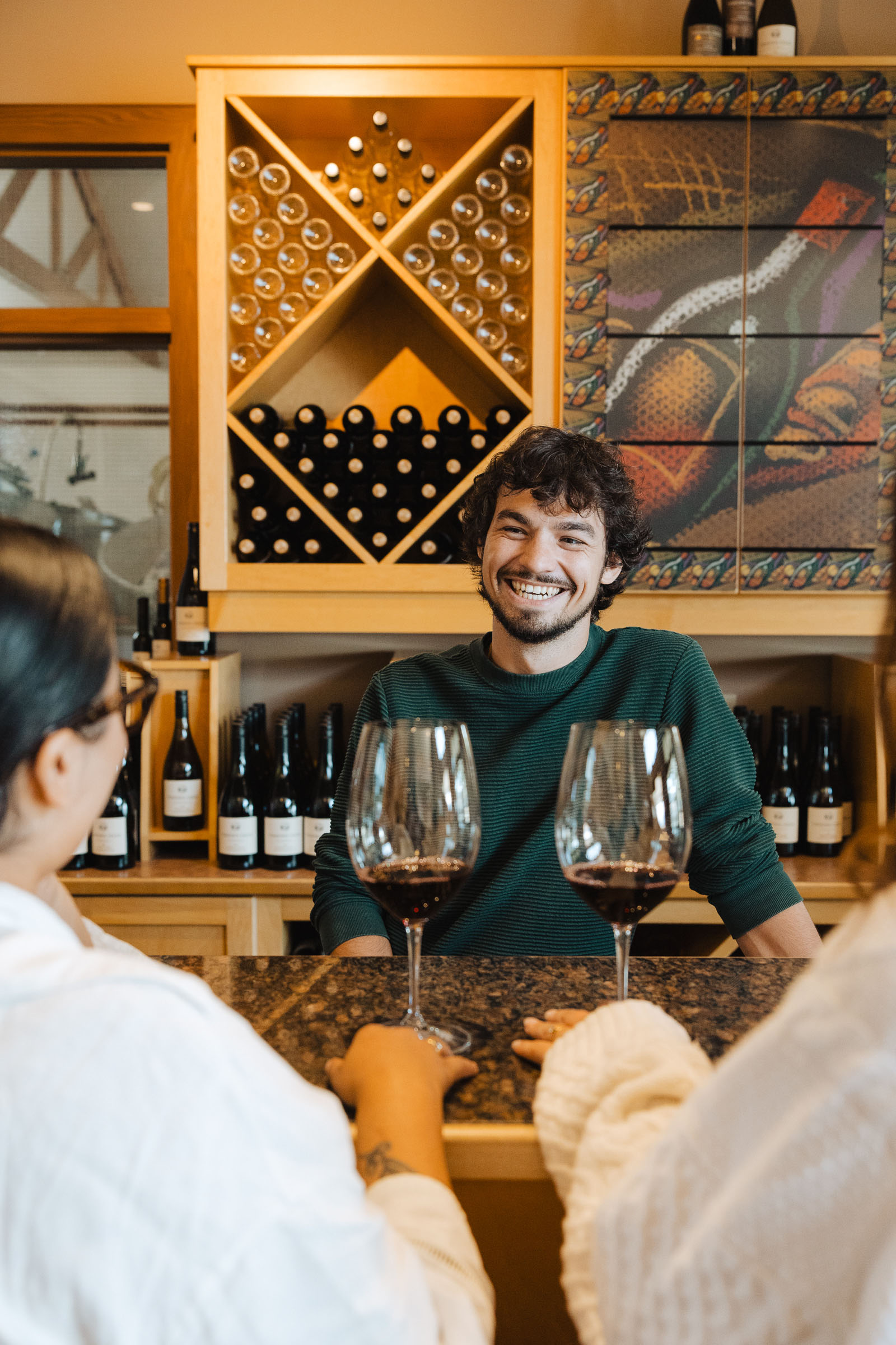 A smiling man stands behind a wine bar with a wine rack in the background. Two women with long hair are seated at the bar, each holding a glass of red wine. The setting is warm and inviting.