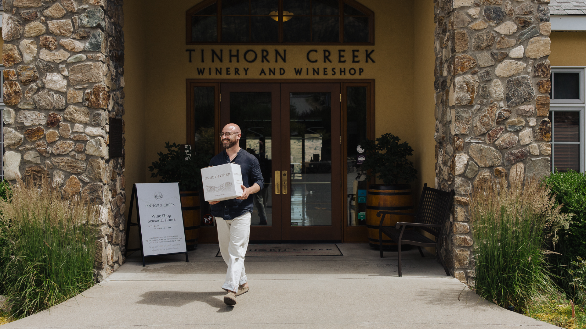 A person carrying a box walks out of the entrance of Tinhorn Creek Winery and Wineshop. The building features a stone archway and glass doors. A sign with information is placed on the left side of the entrance.